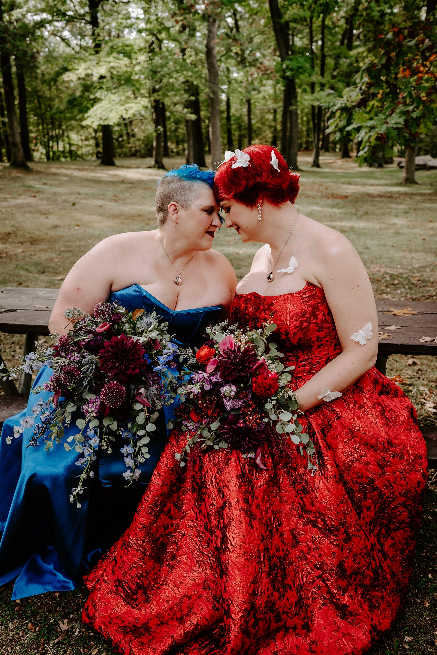 The author and her wife sit at a picnic table, foreheads resting on each others, in red and blue wedding dresses.
