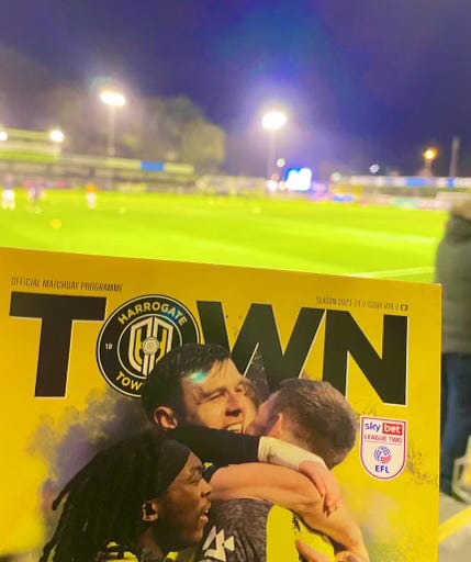 The programme for Harrogate Town vs. Newport with the pitch in the background, the floodlights and a dark sky.