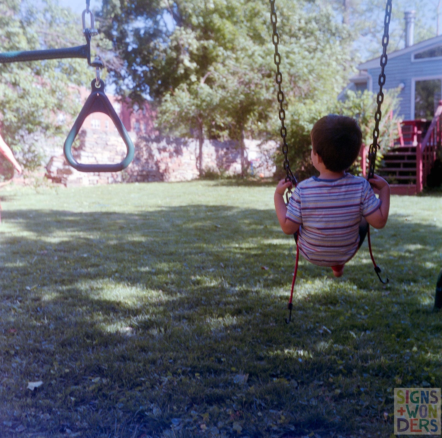ID: The back of a small boy in a striped shirt as he sits on a swing.