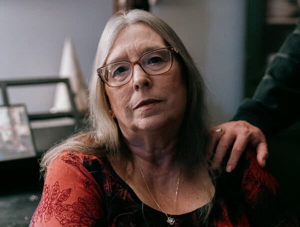 Lydia Birk wears glasses and a red patterned top as she poses for a portrait with her husband's hand on her shoulder.