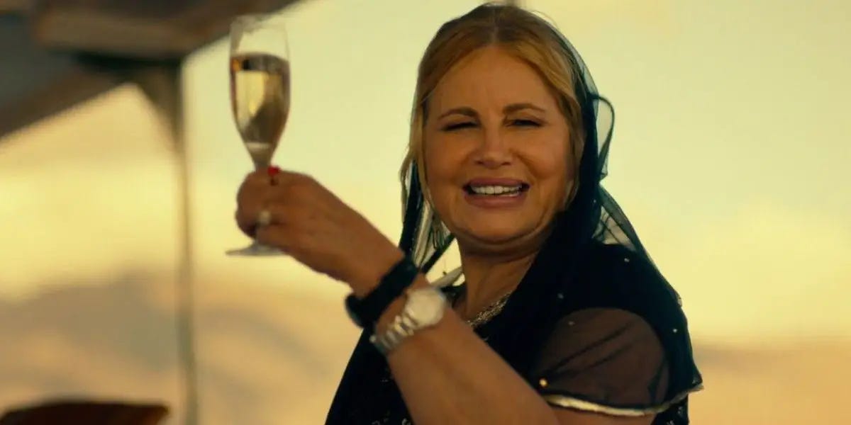 A blonde white woman is wearing a nice black dress with a gauzy shawl. She smiles as she raises a glass of champagne from an open-air boat.