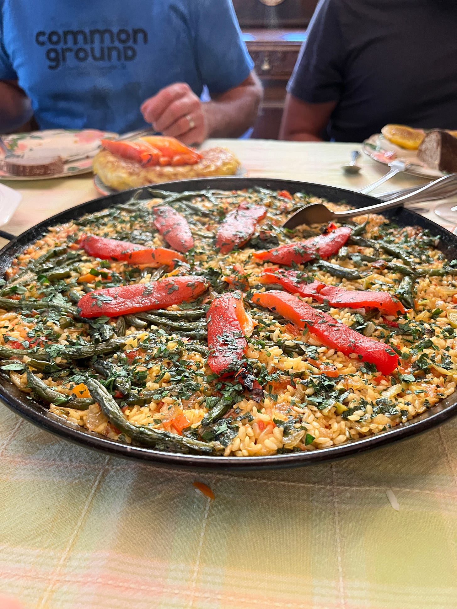 This is an image of the paella on the table. It is in a dark black pan and has a spoon in it. There are bright red veggies atop the paella in a starburst pattern.