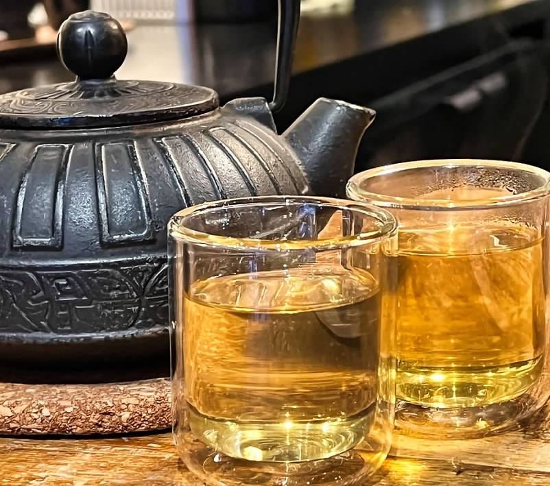 Two glass cups filled with golden tea sit beside a black, ornate cast-iron teapot on a wooden surface