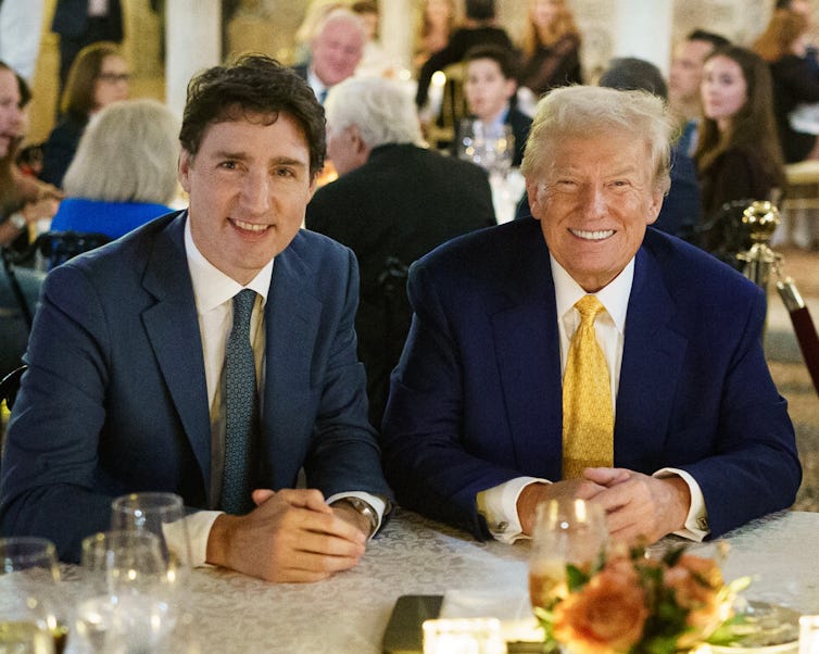 A dark-haired man and an older man with grey-ish blond hair smile at a dinner table.