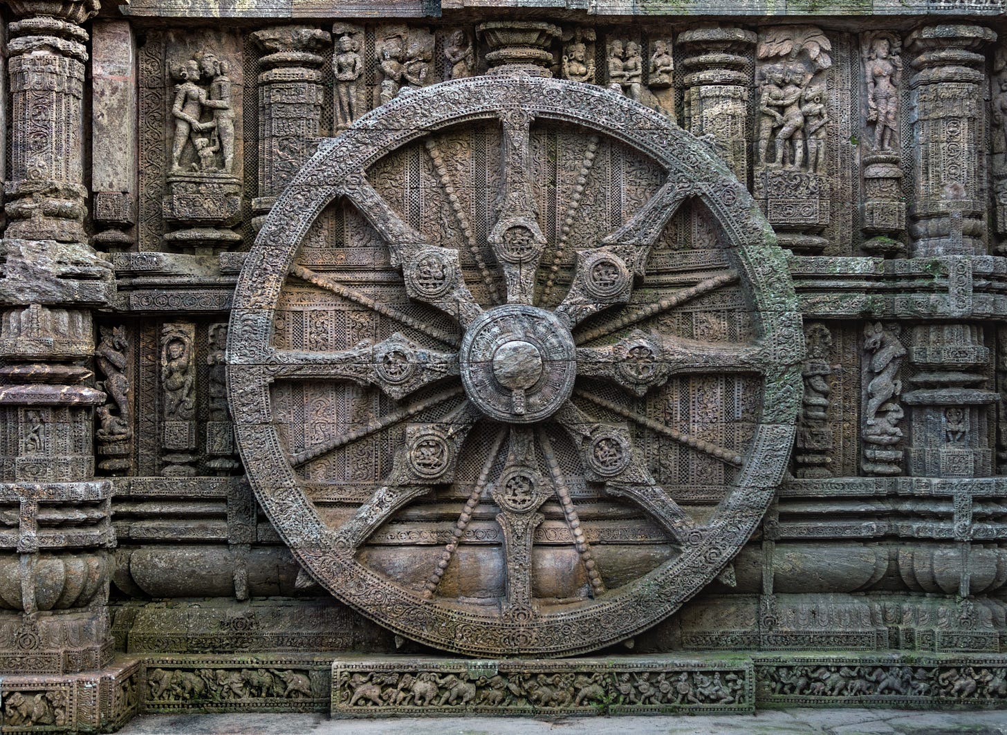 Stone wheel engraving of the Dharmic Wheel. There are eight spokes, a central hub, and an outer wheel. Other engravings adorn the wall: one can make out a border of small elephants along the bottom of the wall.