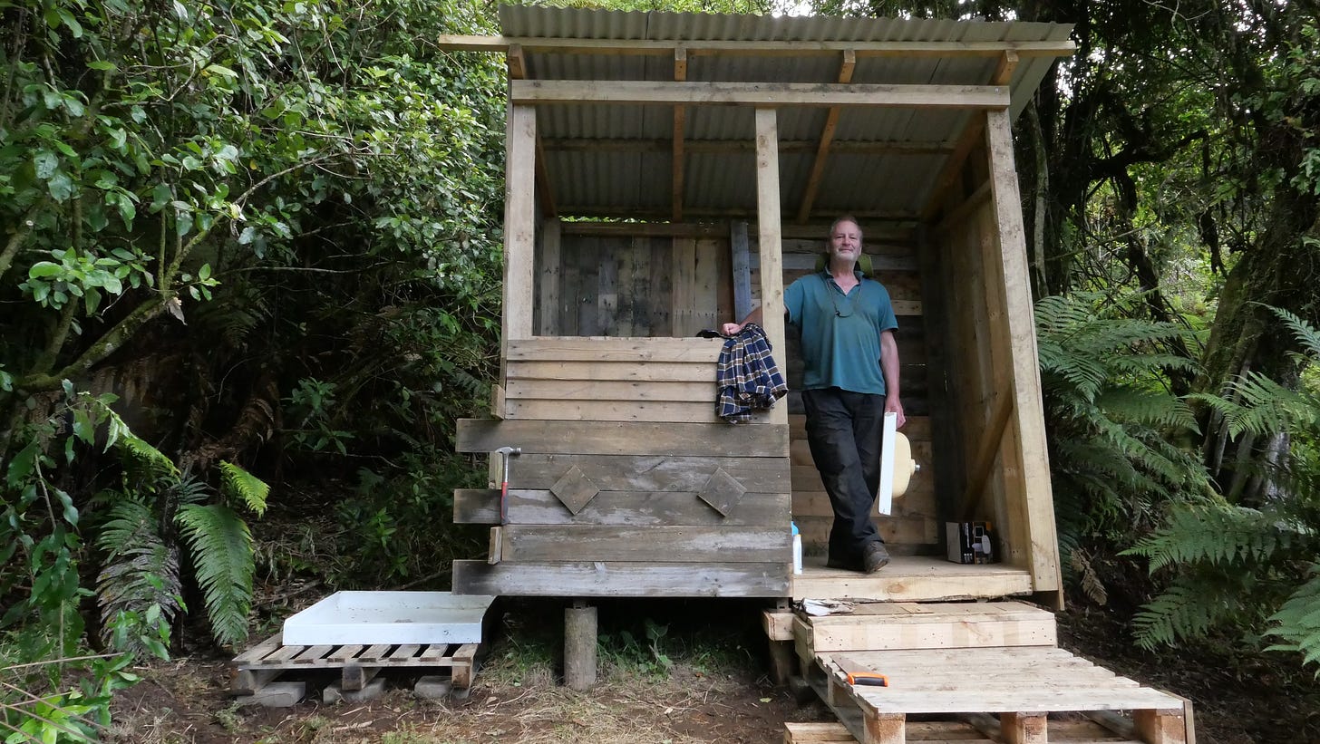 Mike standing in the doorway of a hut that now has wooden walls, a hole for a window, steps and a shower tray to one side.