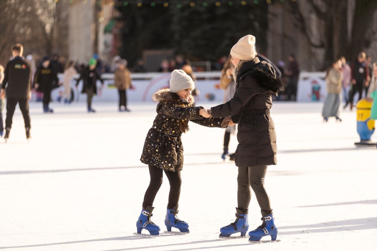 A woman and young girl ice skate together at an outdoor rink