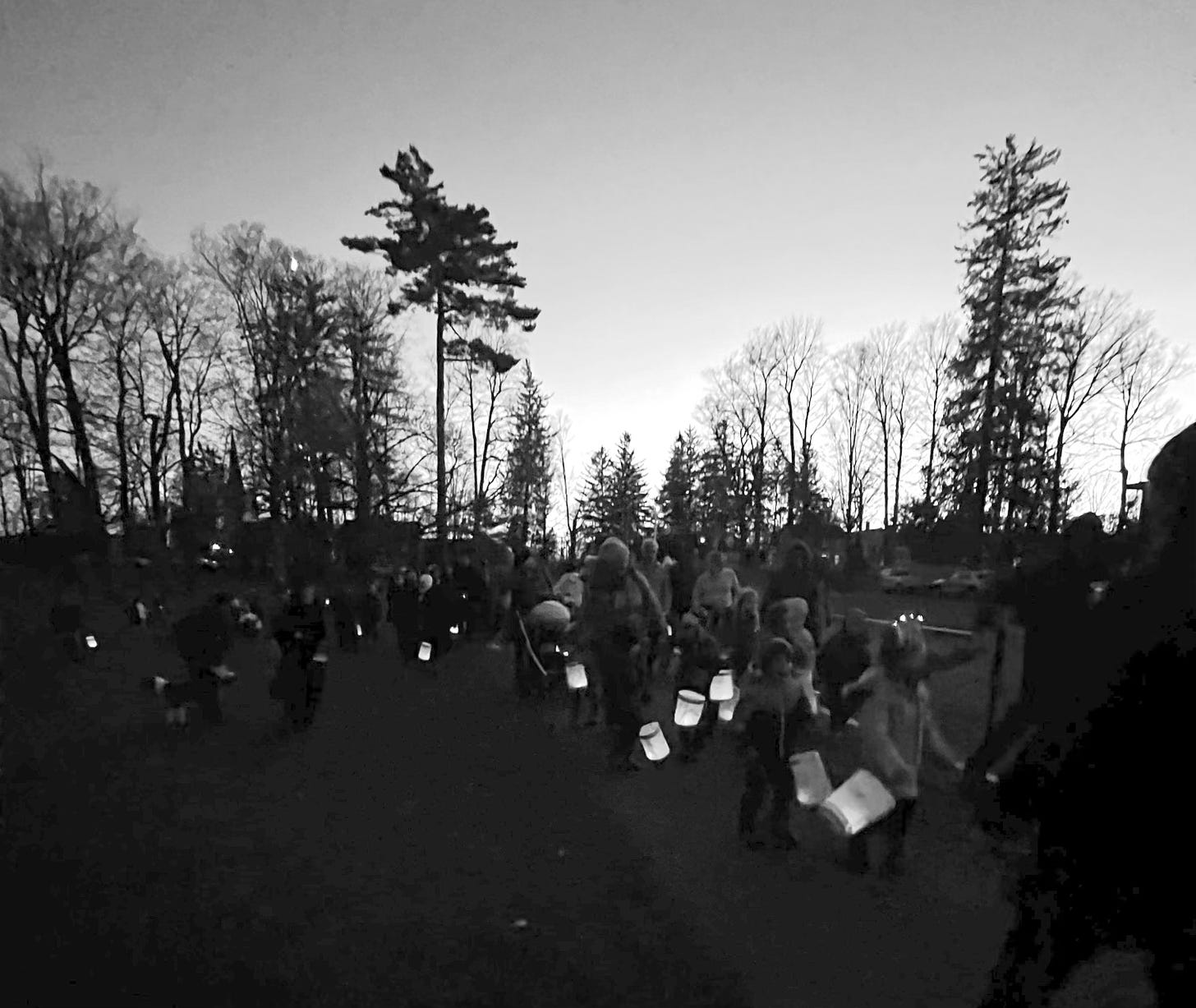 The photograph is grayscale and atmospheric. A parade of children carry paper lanterns at dusk, their shapes like shadows. Their lanterns, bright like fireflies, seem to float. Behind them, a line of trees stands against a darkening sky.