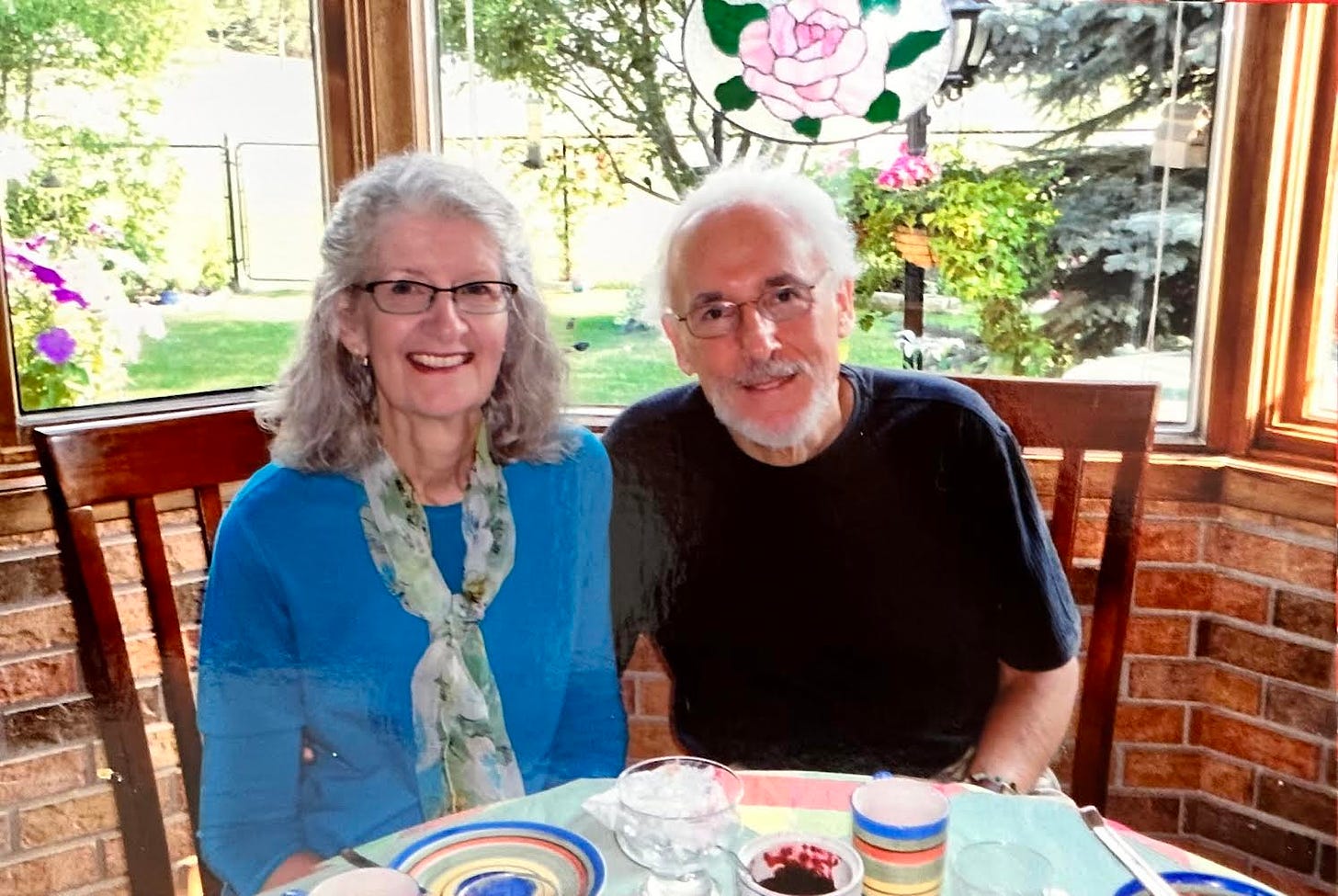 A upper-middle-age woman and man sit at a table, possibly in a restaurant, and smile for the camera.