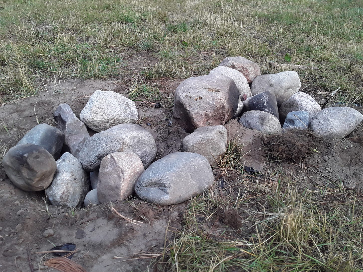 Photograph of two, small, adjacent fire pits dug in to the grass at Davis Street Park, using large rocks that were removed from the skateboard plaza.  Four of the rocks are partially scorched black from fire.