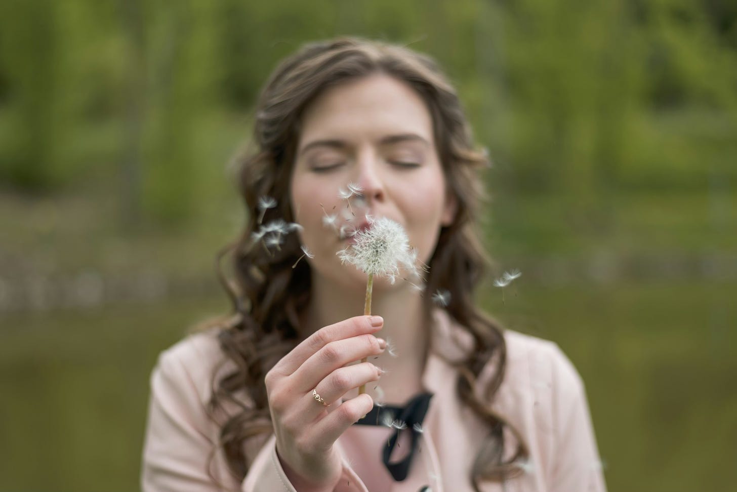 Woman with long hair wearing a pink sweater blows on a dandeloin with her eyes closed.