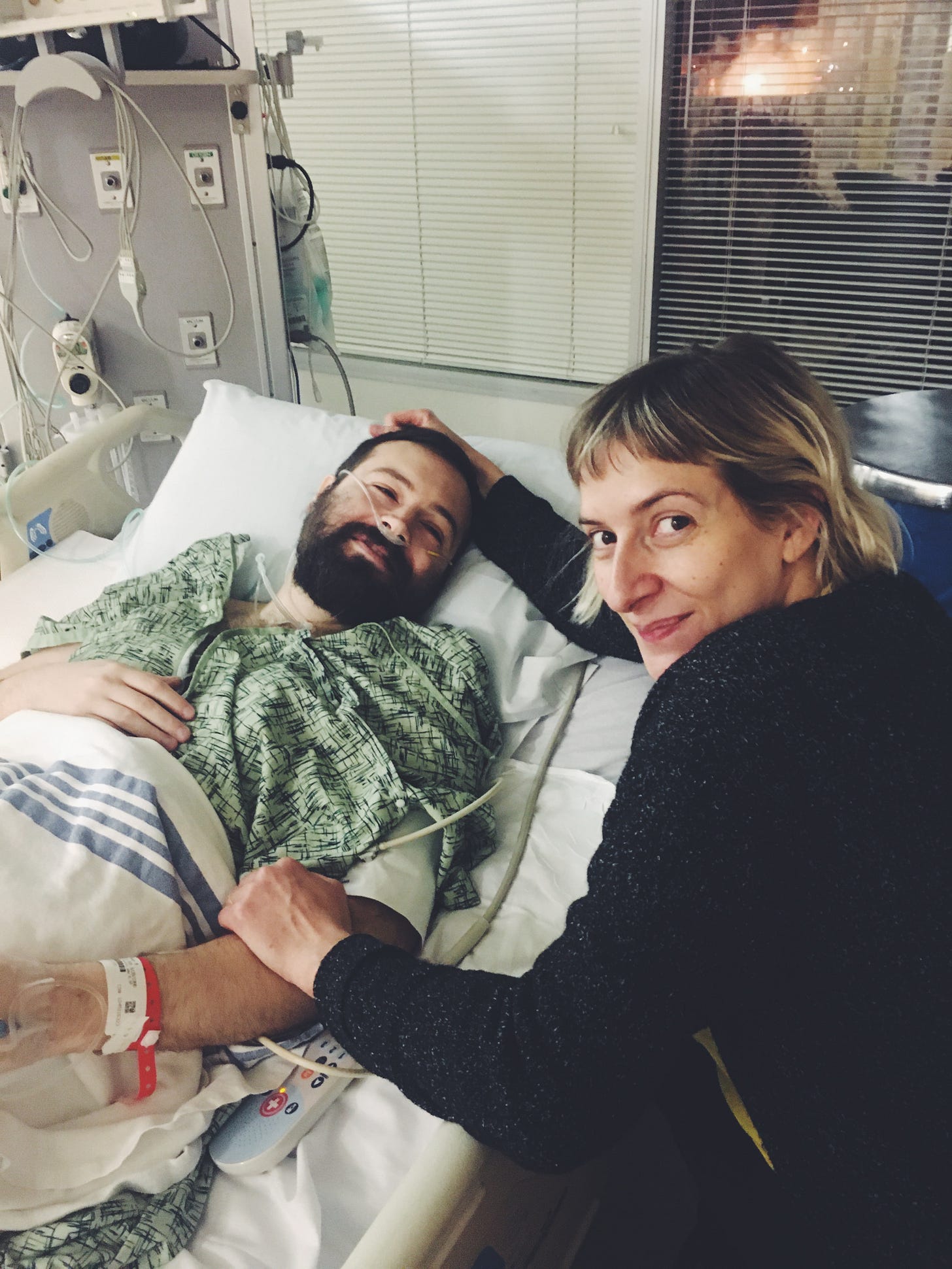 Rebekah sits next to her partner Micah who is laying in a hospital bed recovering from surgery. He is in a green gown with oxygen tubes in his nose, a blood-pressure cuff on his arm, and IV in his hand, and bands on his wrist. Rebekah has one hand around his arm, the other on the top of his head. Rebekah sits next to her partner Micah who is laying in a hospital bed recovering from surgery. He is in a green gown with oxygen tubes in his nose, a blood-pressure cuff on his arm, and IV in his hand, and bands on his wrist. Rebekah has one hand around his arm, the other on the top of his head.