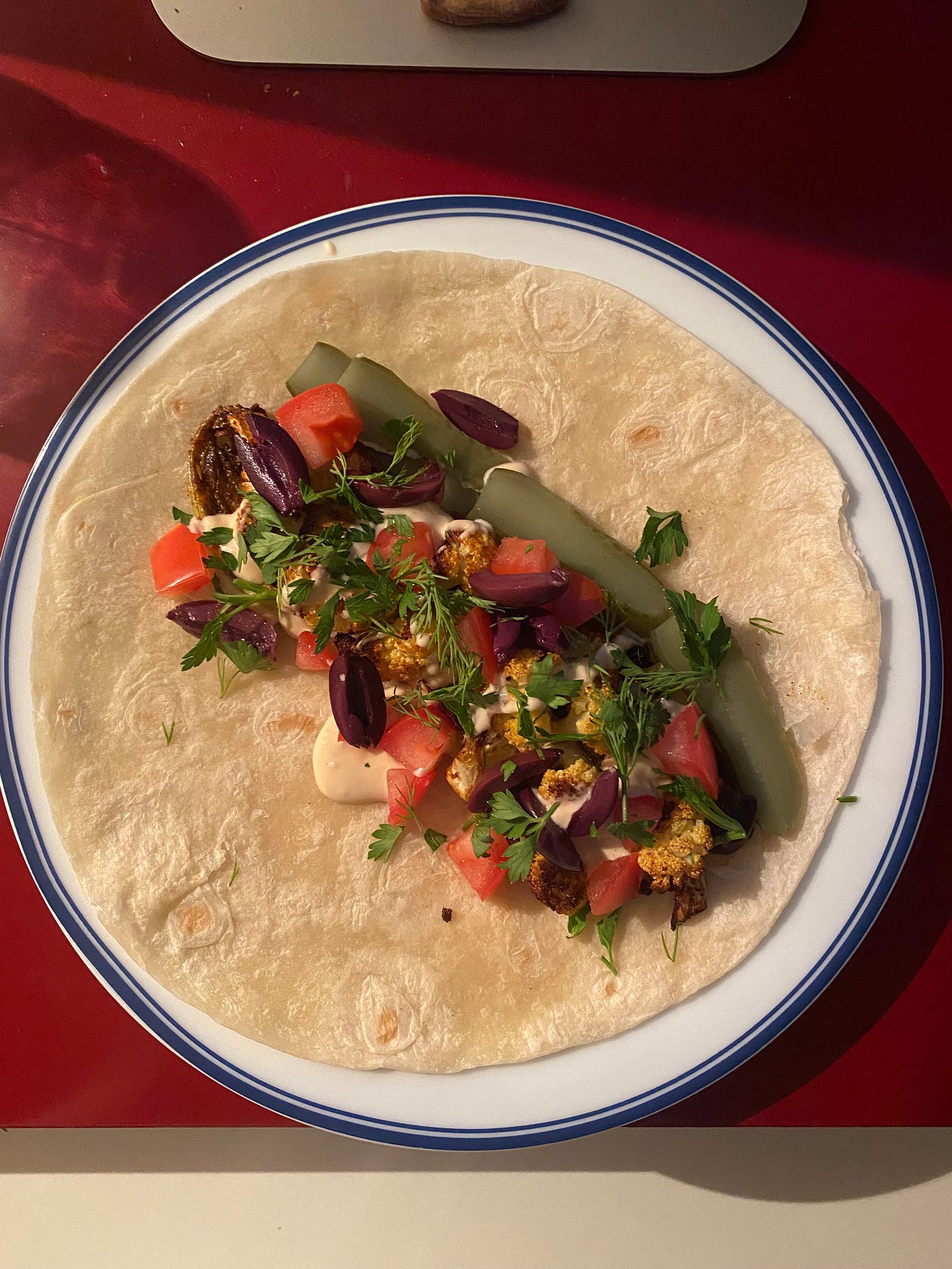 A white plate with a blue rim on top of a red desk. On the plate is an open tortilla with the roasted cauliflower shawarma described above, topped with tahini sauce, kalamata olives, pickles, diced tomato, and herbs.