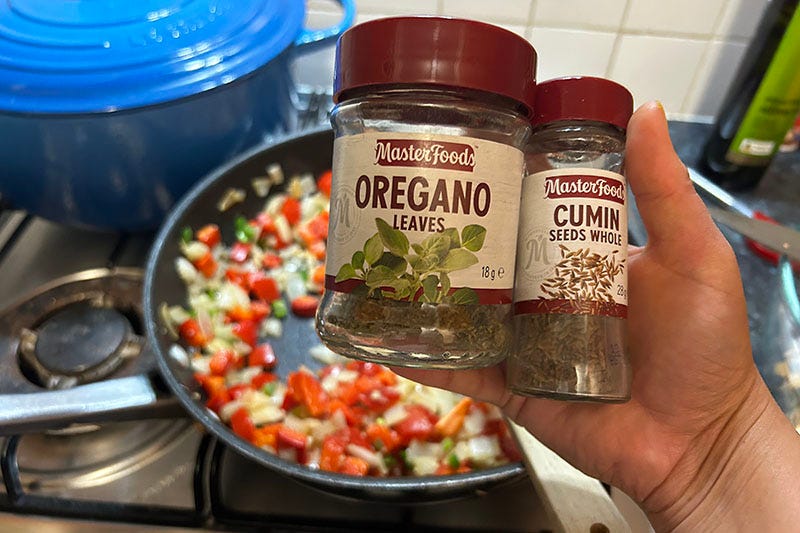 A hand holds jars of oregano and cumin seeds above a frying pan full of veg.