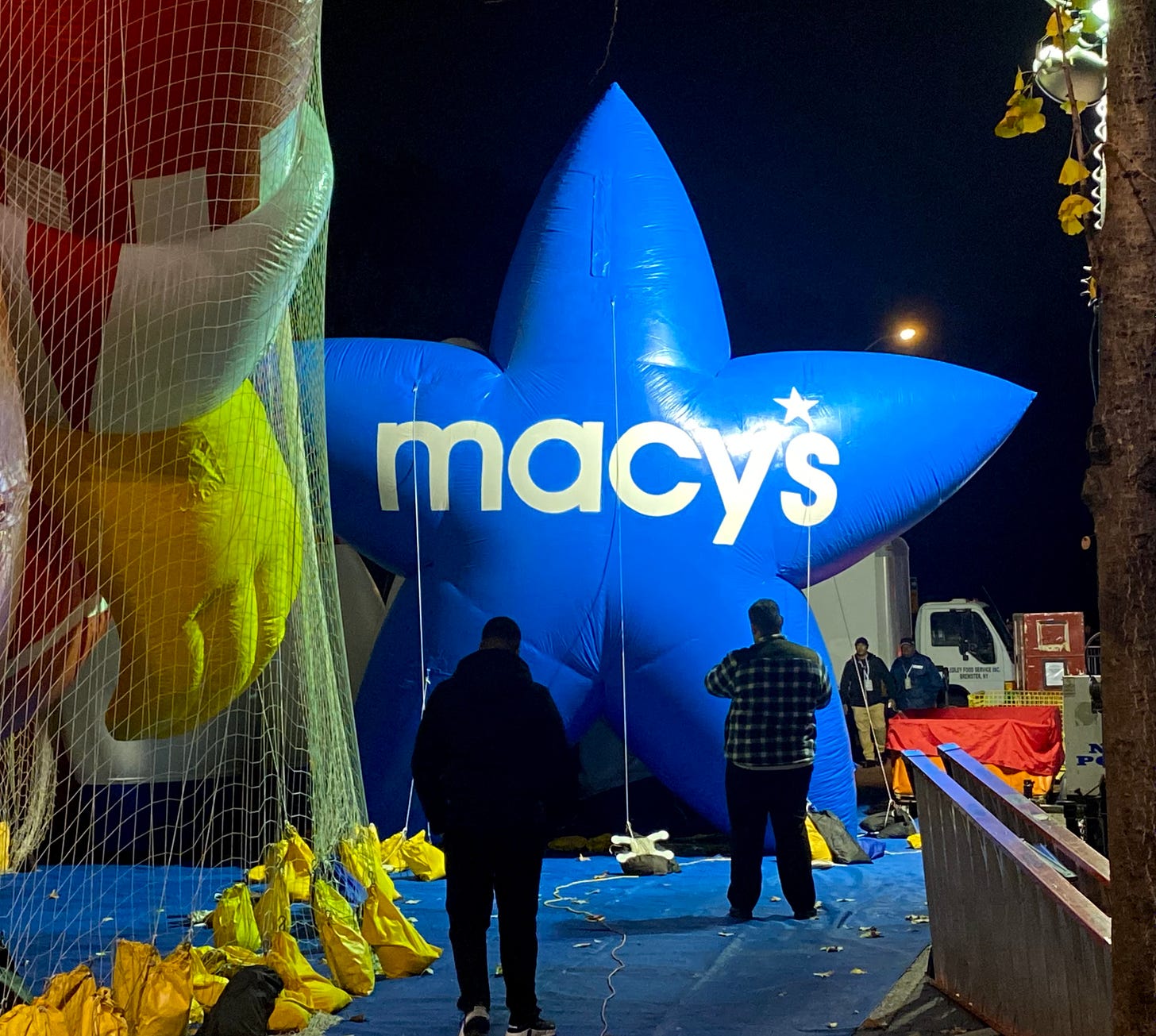 A blue star parade ballon with Macy's written on it in white letters, sandbagged next to a Ronald McDonald balloon tied down in netting. Two men are in front of it, working.