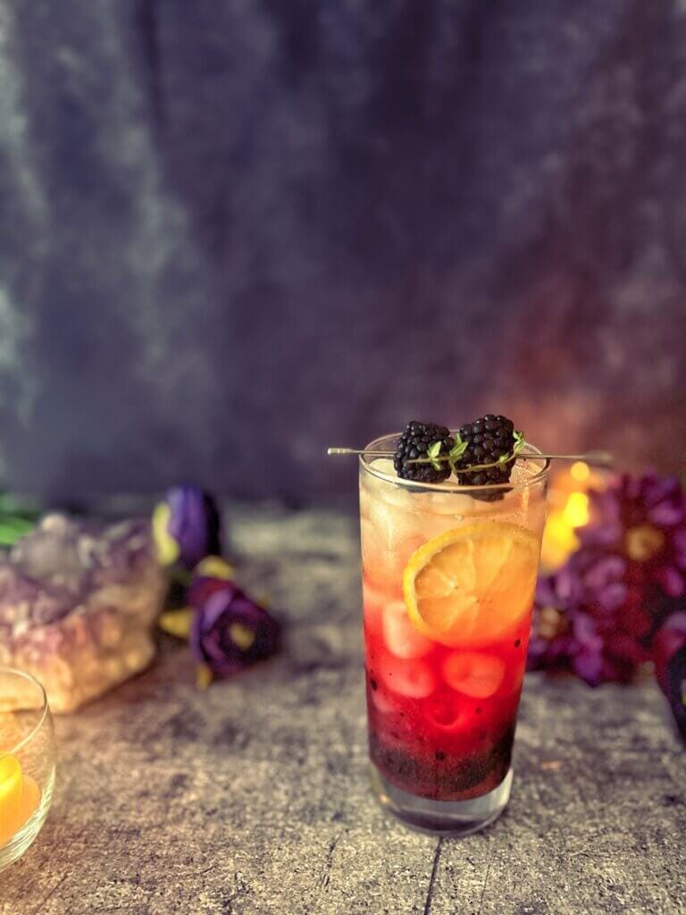 A purple and gold mocktail in a tall skinny glass, garnished with a lemon wheel and skewered blackberries