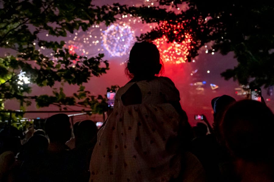 A girl sits on someone's shoulders, between trees, as fireworks explode in the distance.