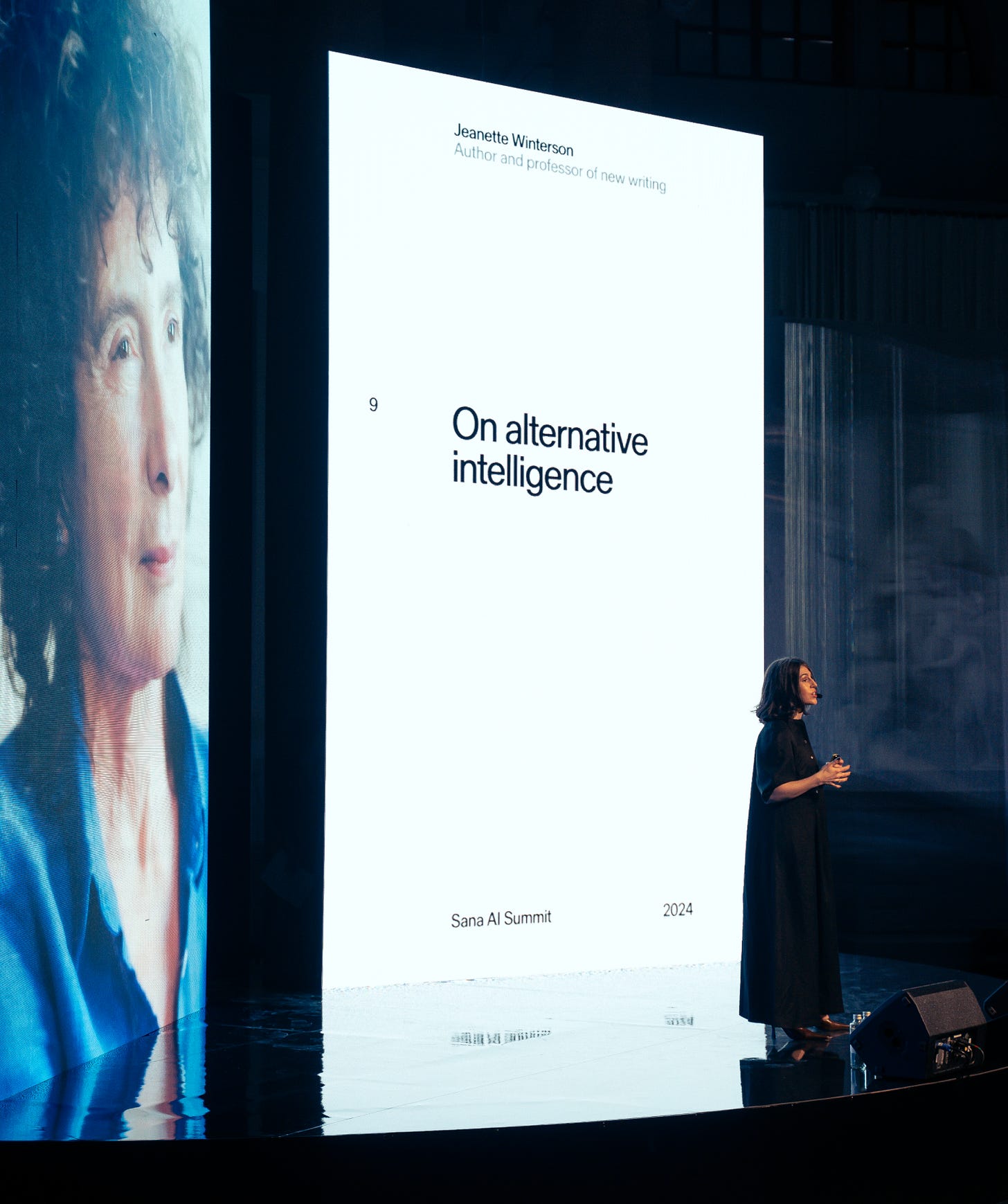 Photograph of Pass It On creator Lauren Crichton standing in front of two giant LED screens on stage at Sana AI Summit. On one LED screen is a photograph of Jeanette Winterson. On the other reads the title of Jeanette's keynote "On alternative intelligence."