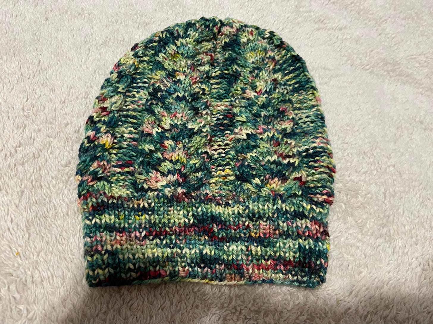 A cable hat knitted in variegated greens and reds laying on a cream-colored throw blanket.