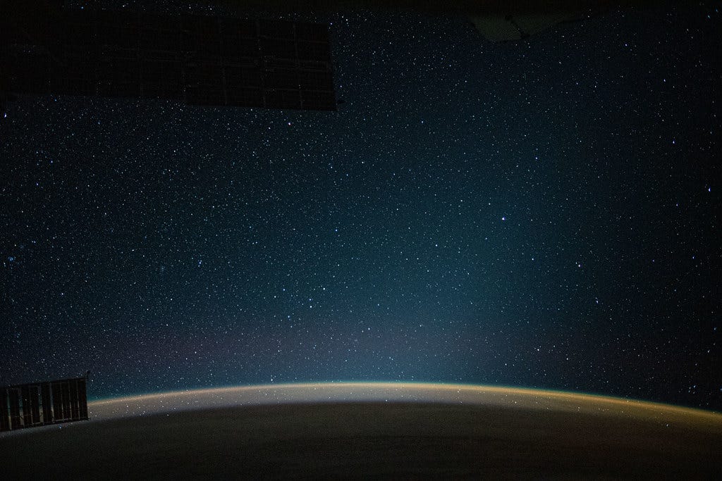 a starry night sky above the curving horizon "An atmospheric glow blankets Earth beneath a starry night sky" by NASA Johnson is licensed under CC BY-NC-ND 2.0.