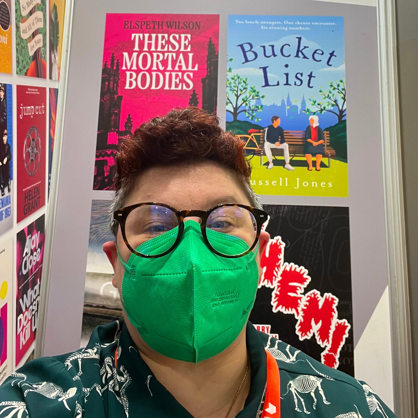 A white person with short red hair and glasses and a green face mask. They are wearing a green shirt with dinosaurs on and a red lanyard and are sitting in front of a book fair panel