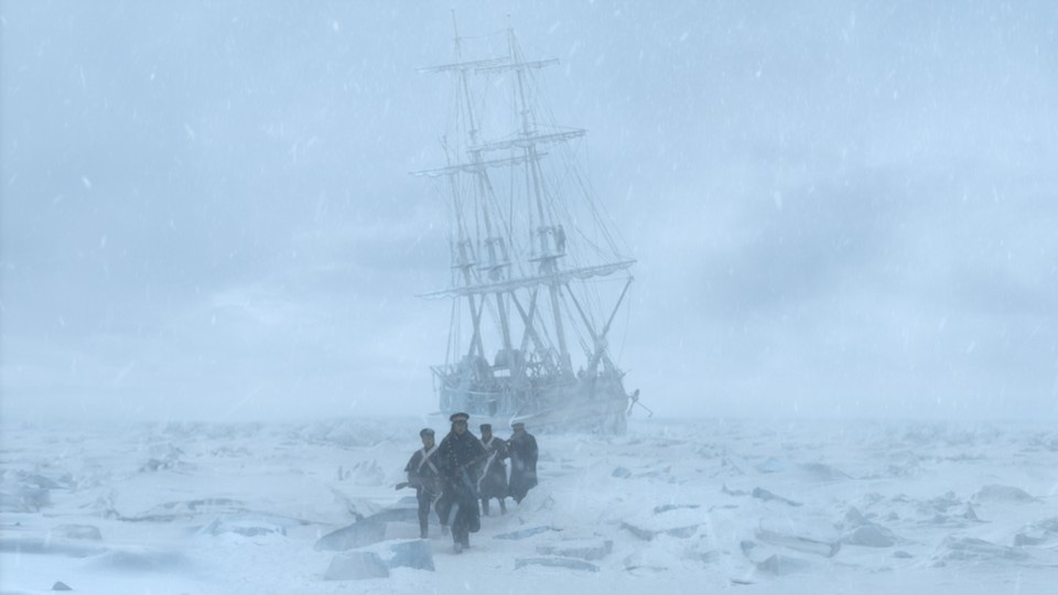 A small group of men walk across a snow-covered landscape toward the camera and away from an old ship.