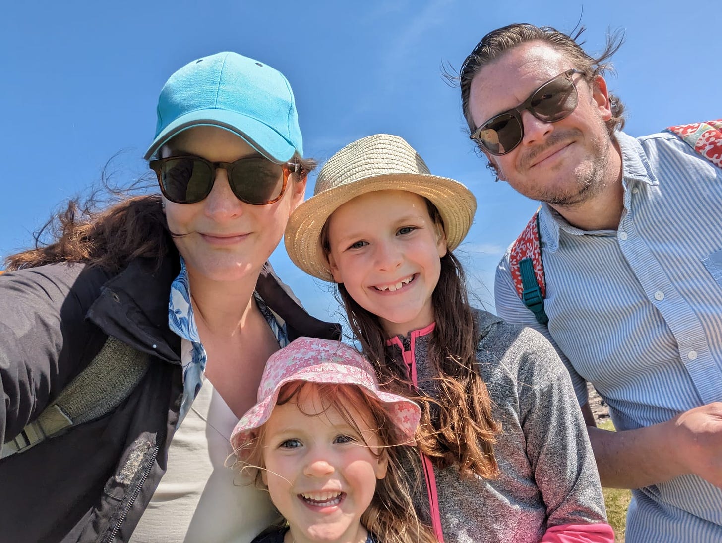 A family of four, all white with brown hair. Mel on the left has a blue baseball cap she nicked from her eldest. The two children have hats on and big toothy grins. The two adults have more of a grin and bear it closed-lip smile. 