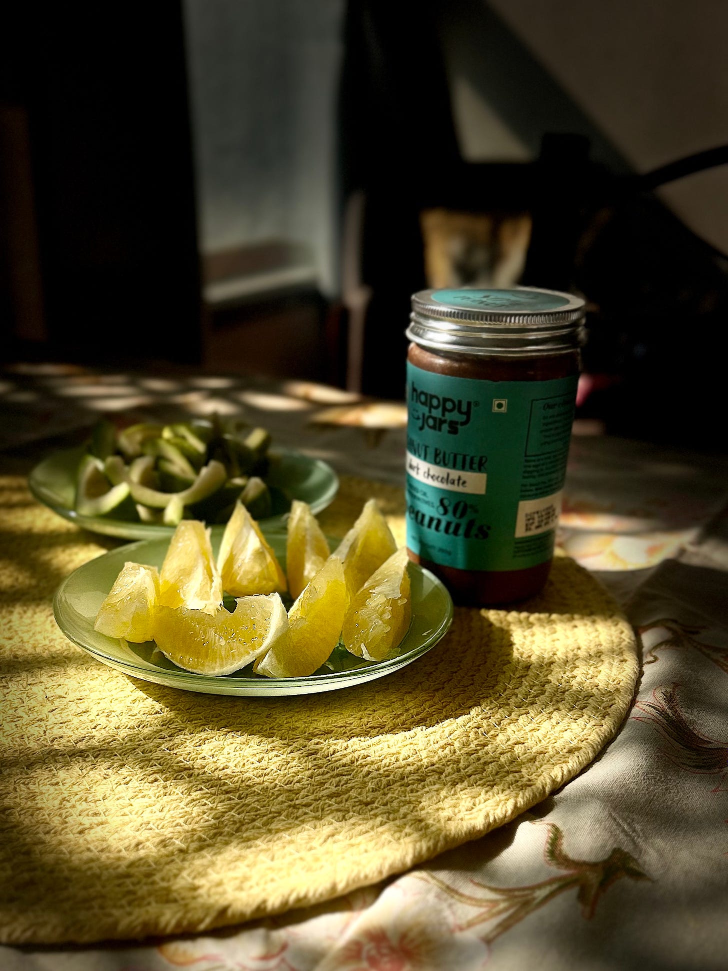 A beautiful picture of sun falling on a dining table in the morning hours. The yellow and green of the fruits and a peanut jar compliments the sunshine coming in the room.