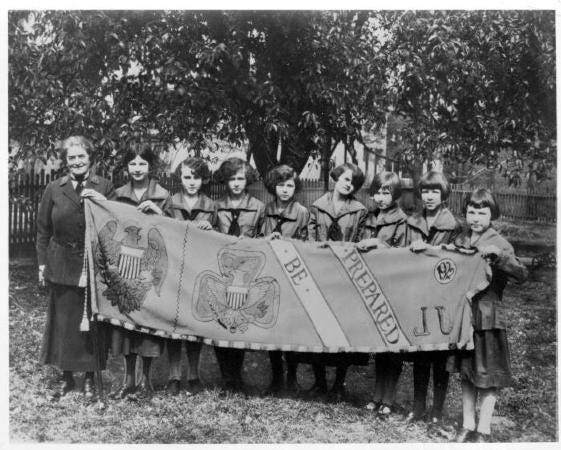 Juliette Gordon Low in uniform poses with Girl Scout troop that holds banner in front of them depicting Girl Scout symbols and slogan.