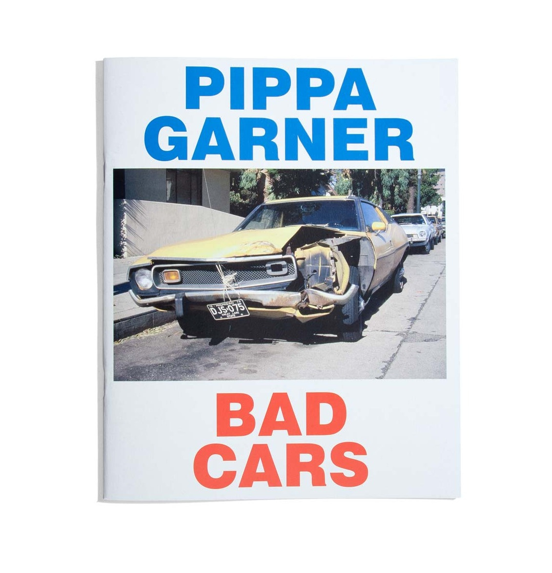 The front cover of Bad Cars by Pippa Garner, a 32 page booklet containing photographs of badly repaired vehicles encounted on the streets of Los Angeles. The front cover design includes a photo of a pale yellow, beat up 70s or 80s car model. The artist's name, Pippa Garner, is in bold blue lettering at the top, and the title "Bad Cars" is at the bottom in bold red lettering.