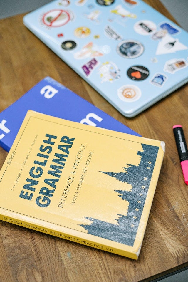 A photo of an English Grammar book, on top of another blue book, and next to a tablet and a highlighter, sat on a wooden table.