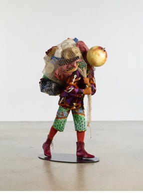 The image depicts a museum sculpture. A mannequin of a boy is dressed in colorful garments in West African patterns, but the clothes themselves are vaguely Victorian and formal. The boy's head is a globe. He carries a fishing net slung over his shoulders. The net is filled with garbage, mostly plastic and styrofoam. The boy is in a walking position, his head tilted downward, seemingly straining against his burden. 