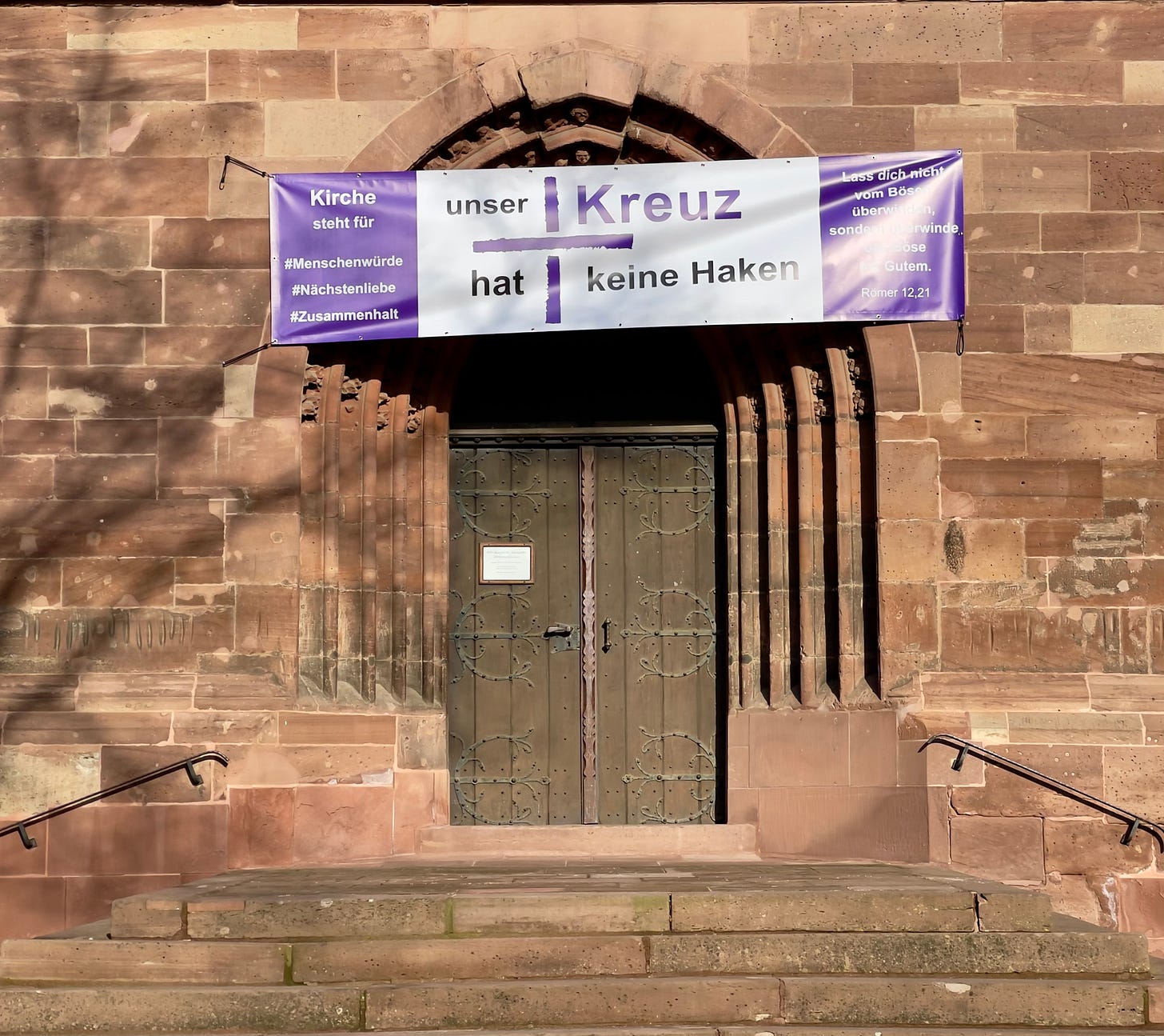 Close-up view of the main entrance of the Münsterkirche St. Alexandri in Einbeck, Germany. A banner above the closed wooden doors reads: “Unser Kreuz hat keine Haken” (“Our cross has no hooks”), with hashtags for human dignity, neighborly love, and solidarity. The church’s late Gothic architecture is visible in the arched doorway. Close-up view of the main entrance of the Münsterkirche St. Alexandri in Einbeck, Germany. A banner above the closed wooden doors reads: “Unser Kreuz hat keine Haken” (“Our cross has no hooks”), with hashtags for human dignity, neighborly love, and solidarity. The church’s late Gothic architecture is visible in the arched doorway.