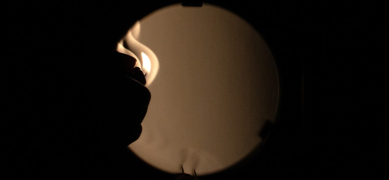 A photograph made in a two mirror Schlieren system with knife edge shows the silhouette of a hand holding a small lit flame in front of a round mirror, staged in a black surround. Typically invisible, heat waves are visible along the golden shadowy gradient of a telescope mirror. At the bottom a candle’s wick and its mirror image break the circle.