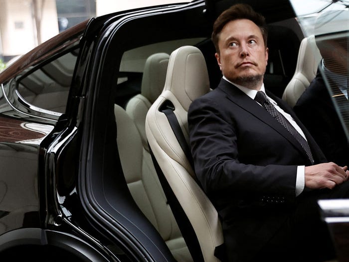 Elon Musk sitting in the a passenger seat of a black Tesla with a white interior while wearing a black suit and tie.