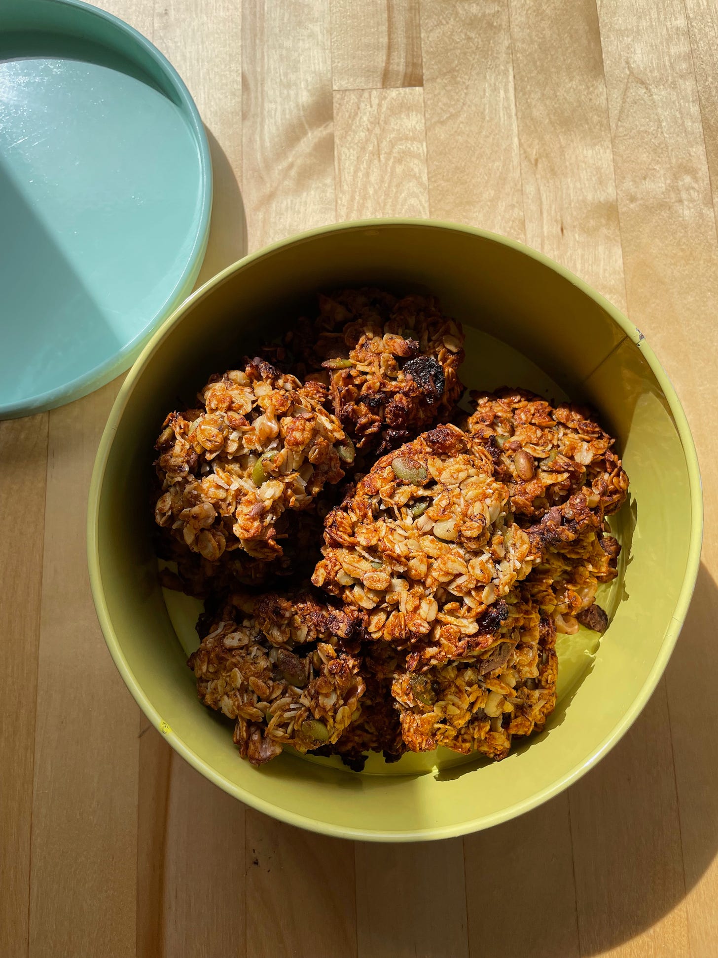 A yellow cookie tin in the sun with golden brown granola cookies inside with pepitas, sultanas and chunks of chocolate. The aqua lid to the round tin in the background.