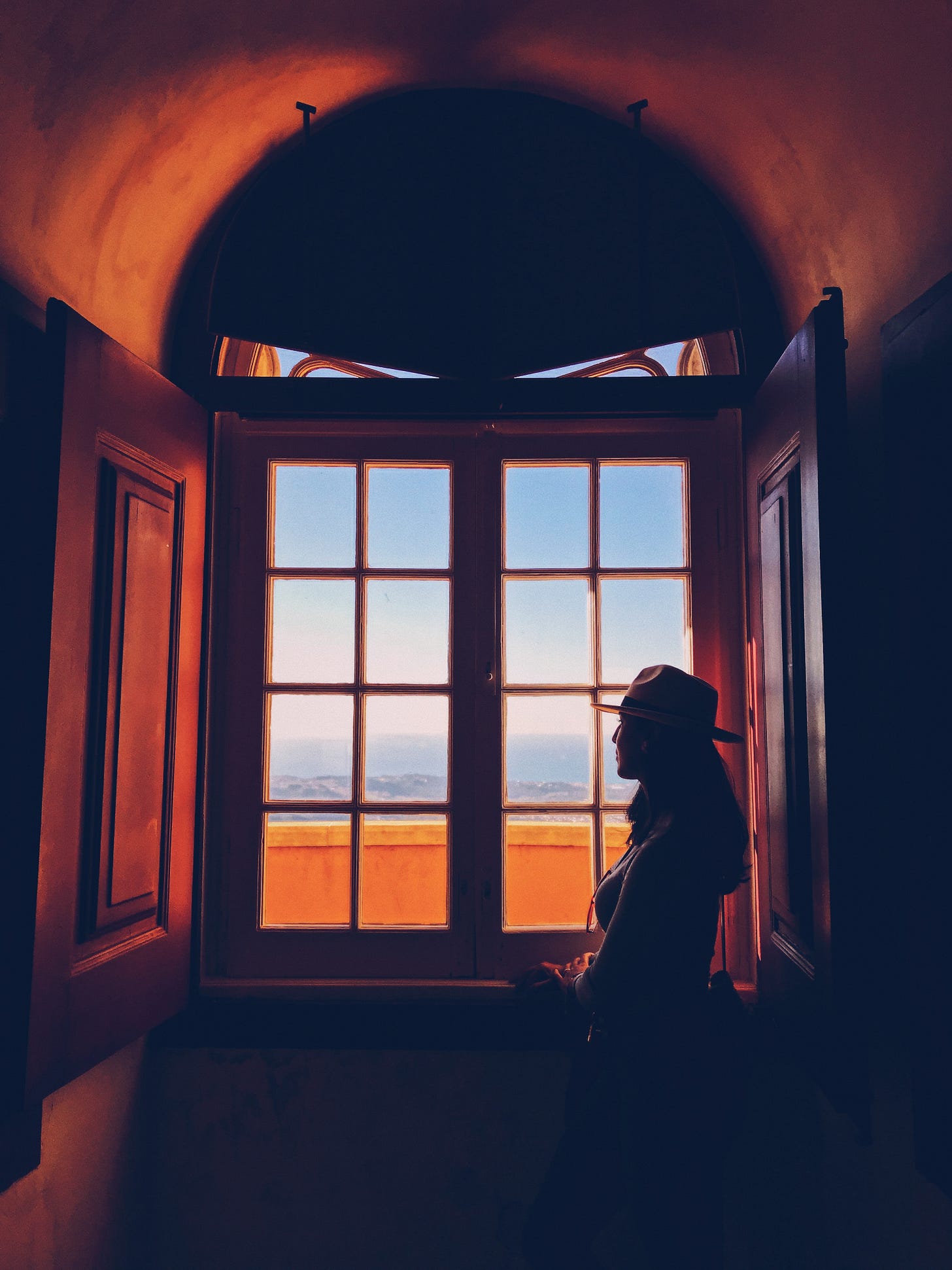 Silhouette of woman wearing rimmed had in red-toned room gazing out paneled window