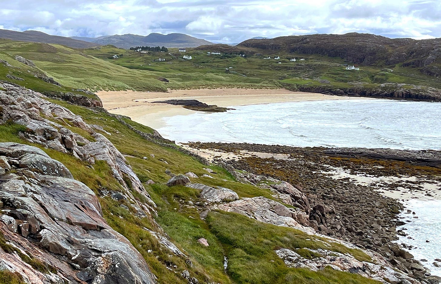 rocky foreground with mossy green grass, calm gray sea and curve of pinkish sand, small white houses in the low hills behind