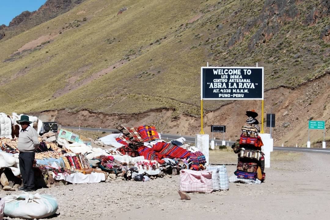 An artisan market in the foreground with a sign that says, “Welcome to Les Desea Centro Artesanal “Abra La Raya” Alt. 4338 M.S.N.M. Puno-Peru”. A road and a scrub-covered hill are in the background.