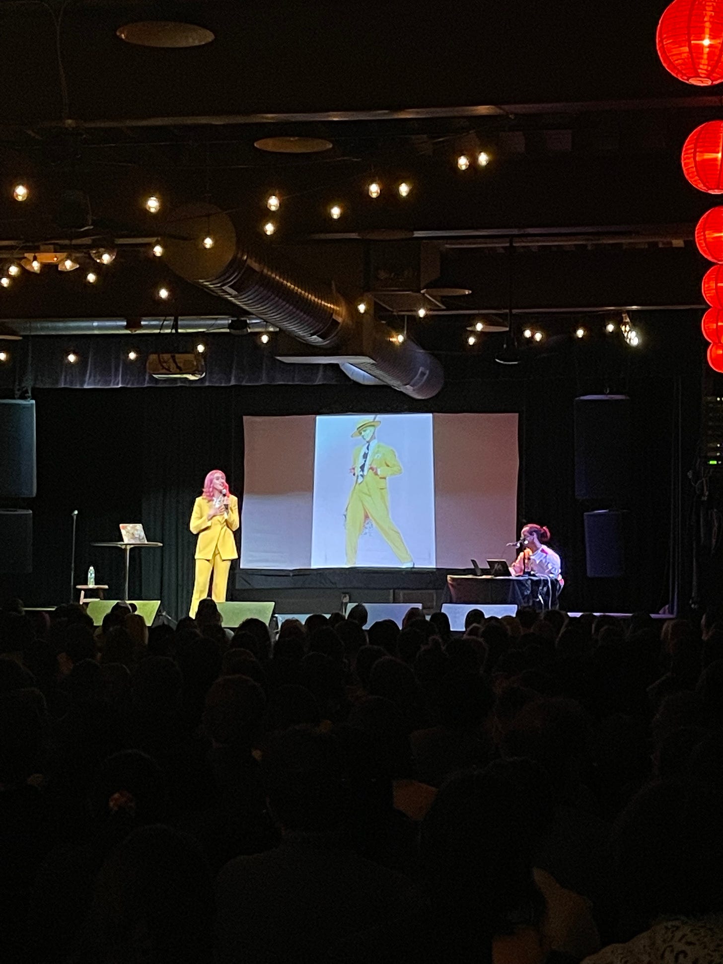 photo of Nora McInerny and Marcel Malekebu on stage at Motorco Music Hall in Durham, North Carolina. Nora is wearing a yellow suit and has hot pink hair