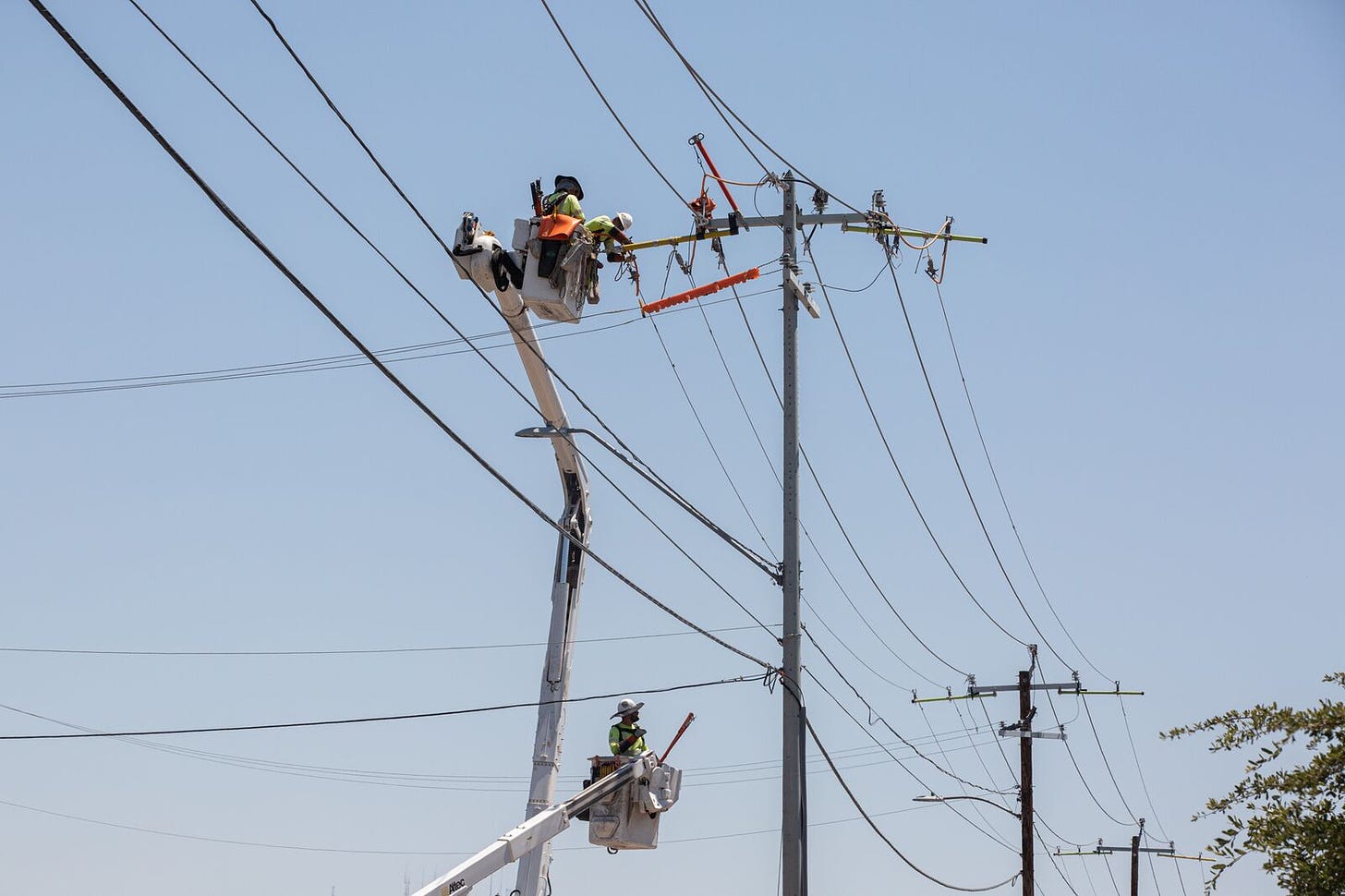 A picture of utility workers in a cherry-picker crane to perform maintenance on power lines. The sky is clear and blue