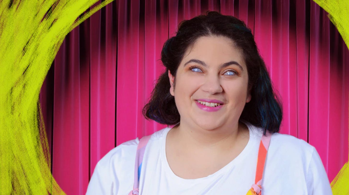 A blind white femme person is smiling, wearing a white shirt and the top of colorful tied overalls, against a brightly lit red velvet curtain and circles of highlighter yellow that seem to be drawn on top of the image.
