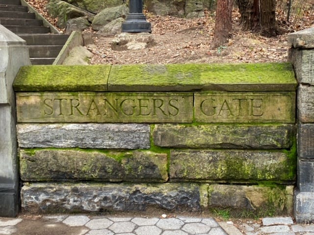 A wall in front of hilly Central Park terrain, next to a entryway leading to stairs. It is engraved, "Strangers Gate."