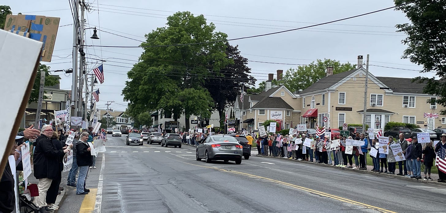A shot down a city street in small-town Maine. A line of protesters stands on the sidewalks as far as the camera can see. Many carry signs. Multiple American flags are in view. A shot down a city street in small-town Maine. A line of protesters stands on the sidewalks as far as the camera can see. Many carry signs. Multiple American flags are in view.