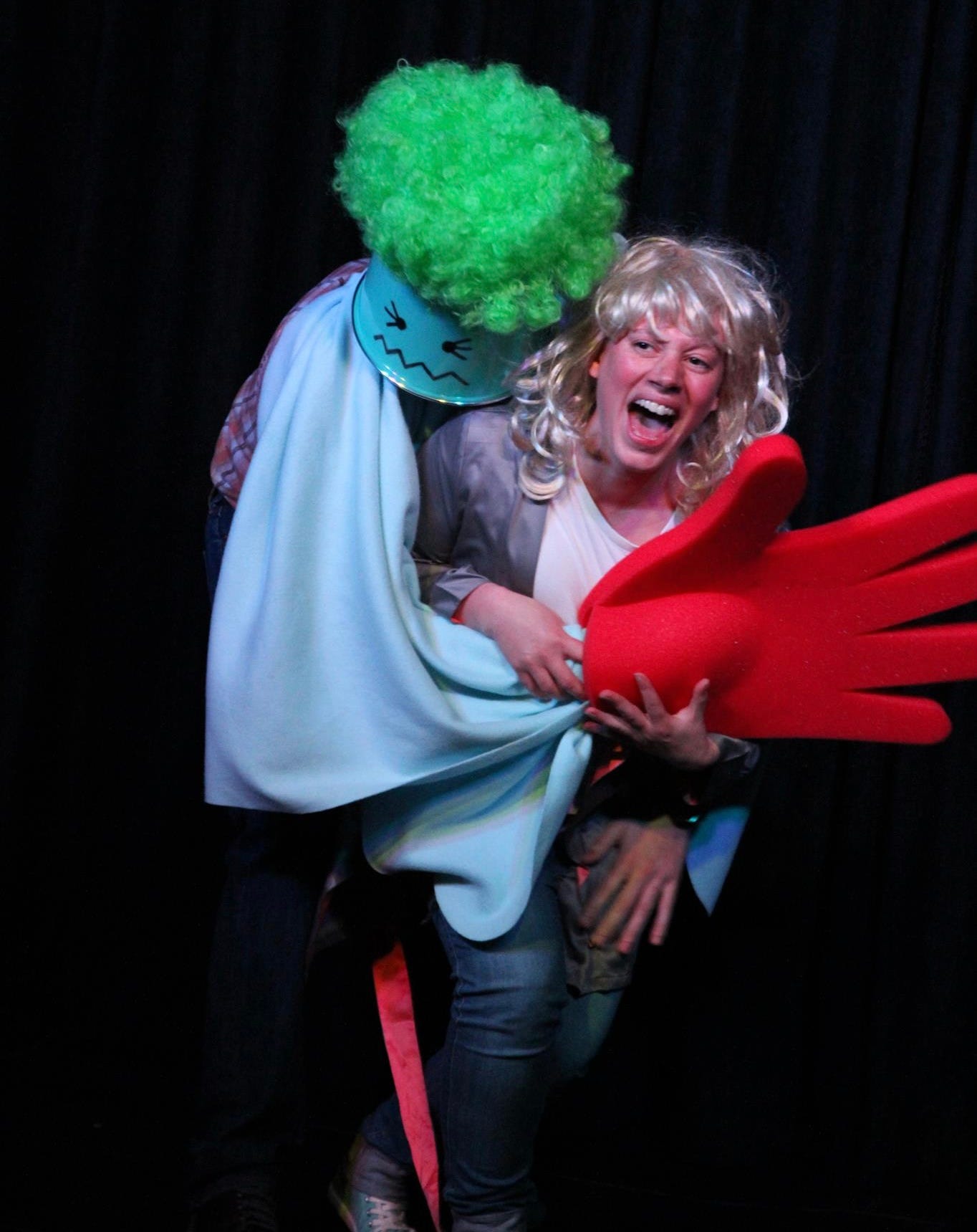 Two performers on stage. One is wearing a blue bin on his head with a face painted on it and a bright green wig. He is clutching the other female performer who wears a blonde wig and is screaming. He envelops her in a blanket and a giant red glove to look like it's his hand