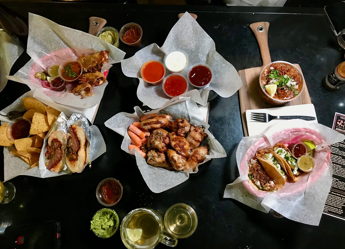 a dining table with chicken wings, a hamburger, tacos and birria with beers