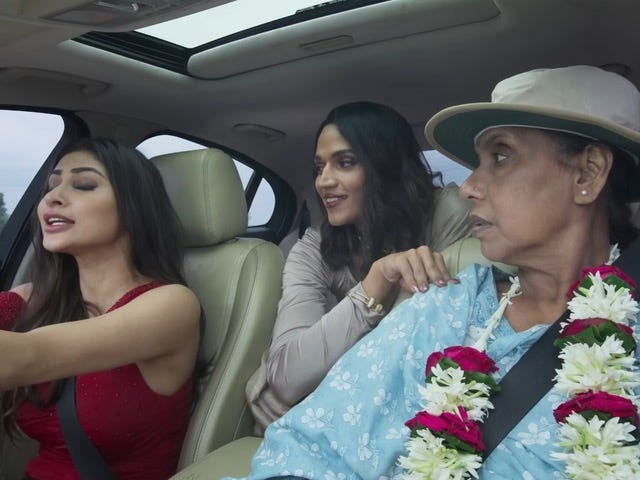 A still from Hindi film, ‘Love Sex aur Dhokha 2 (2024)’. Noor (Paritosh Tiwari) is sitting in a car back seat at the center, leaning forward and smiling as she looks at the lady in the driver’s seat, Mouni Roy who is playing a game show host. The latter is saying something passionately and her eyes are closed. Noor’s mother (Swaroopa Ghosh) is in the passenger’s seat and is looking bewildered at Mouni Roy’s character. Noor is dressed in a beige sari, the game show host is wearing a red sleeveless dress, and the mother is wearing a hat and a blue salwar suit.