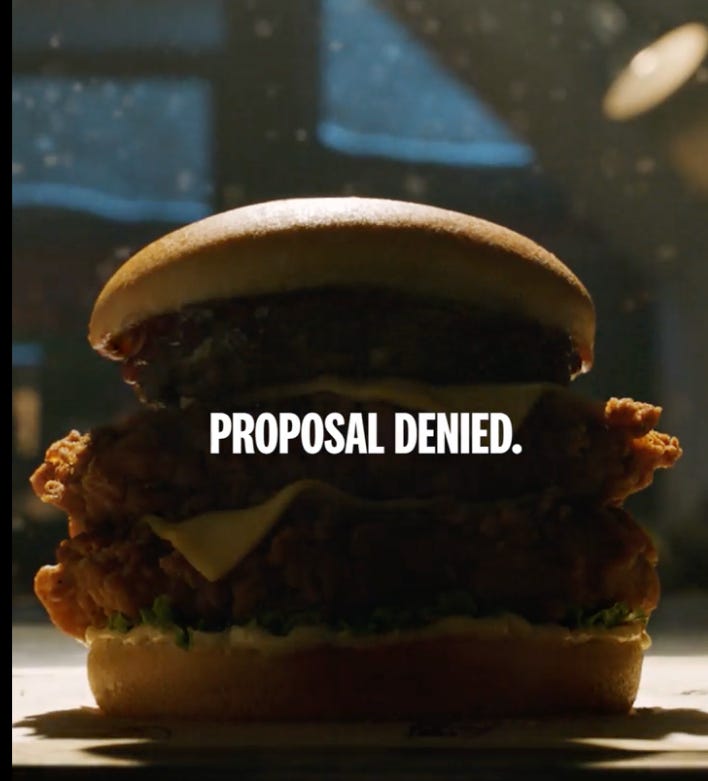 a close-up of a chicken sandwich with the text "PROPOSAL DENIED." prominently displayed over it. The sandwich consists of a crispy chicken fillet, lettuce, and cheese on a bun. The setting appears to be a dimly lit room, possibly a restaurant, with a window in the background showing it's either night or a dark, snowy day.