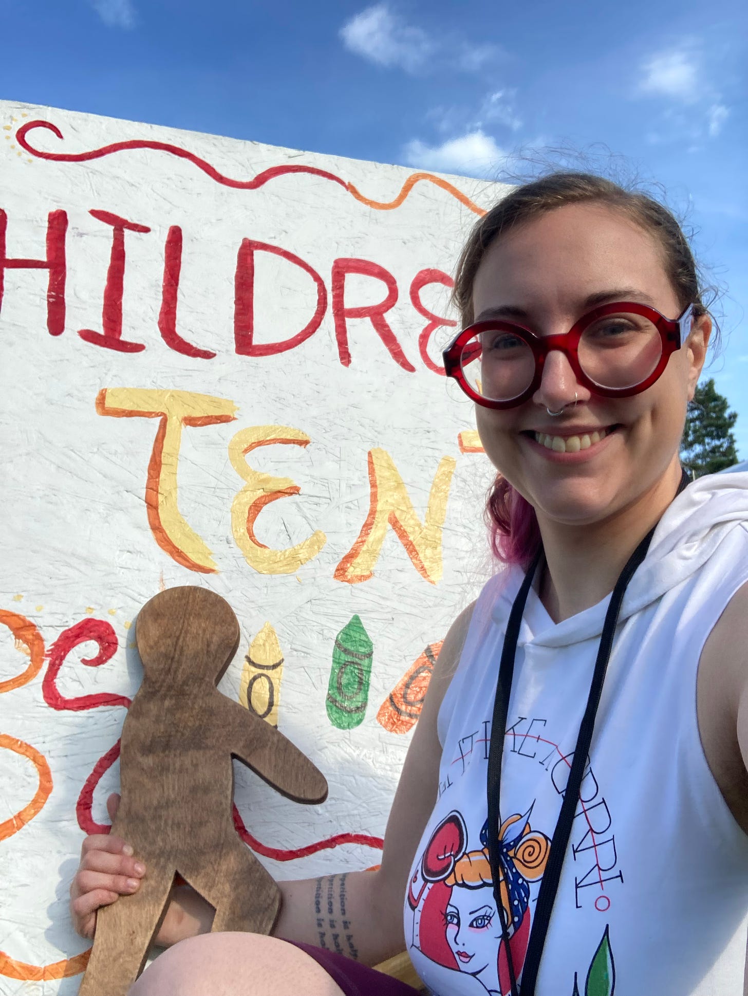 A photo of the author, a white woman with light brown hair and big red glasses, posing with a large wooden person of God in front of a sign reading "Children's Tent"