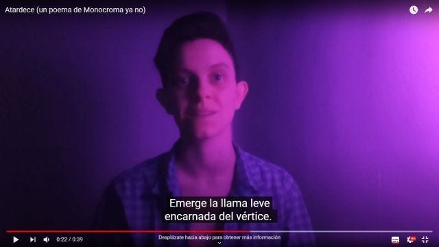 captura de pantalla del vídeo de Miriam leyendo Atardece con los subtítulos Emerge la llama leve encarnada del vértice. Lleva el pelo muy corto y una camisa de cuadros azules y negros. La luz tiene un tono rosa intenso.